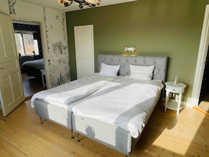 Cama de casal com cabeceira cinzenta e roupa de cama branca, mesa de cabeceira com um vaso, e porta que dá para outro quarto.