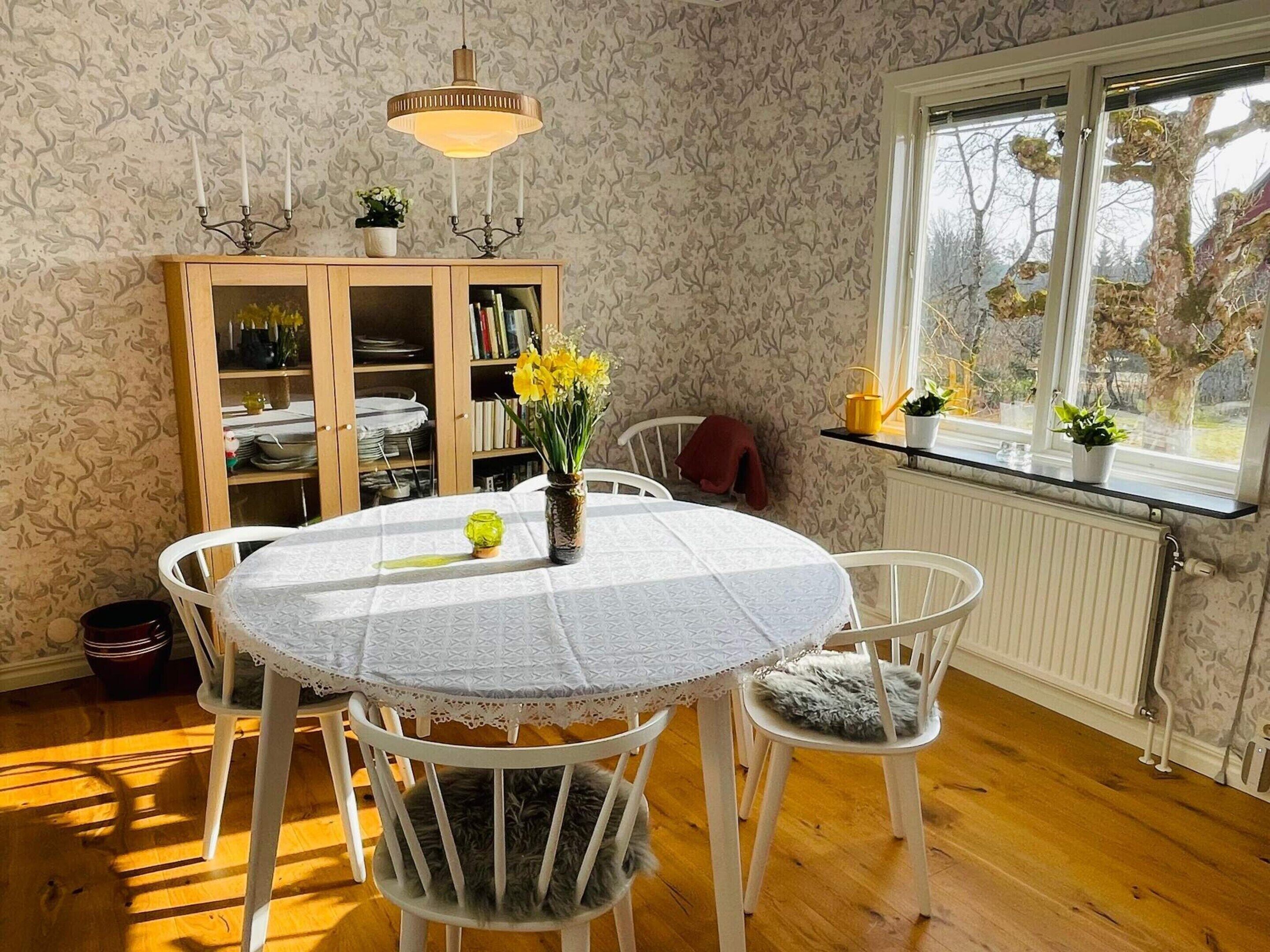 Ein Essbereich mit einem runden Tisch, weißen Stühlen, einem Schrank mit Geschirr und einem Fenster mit Blick auf Bäume.
