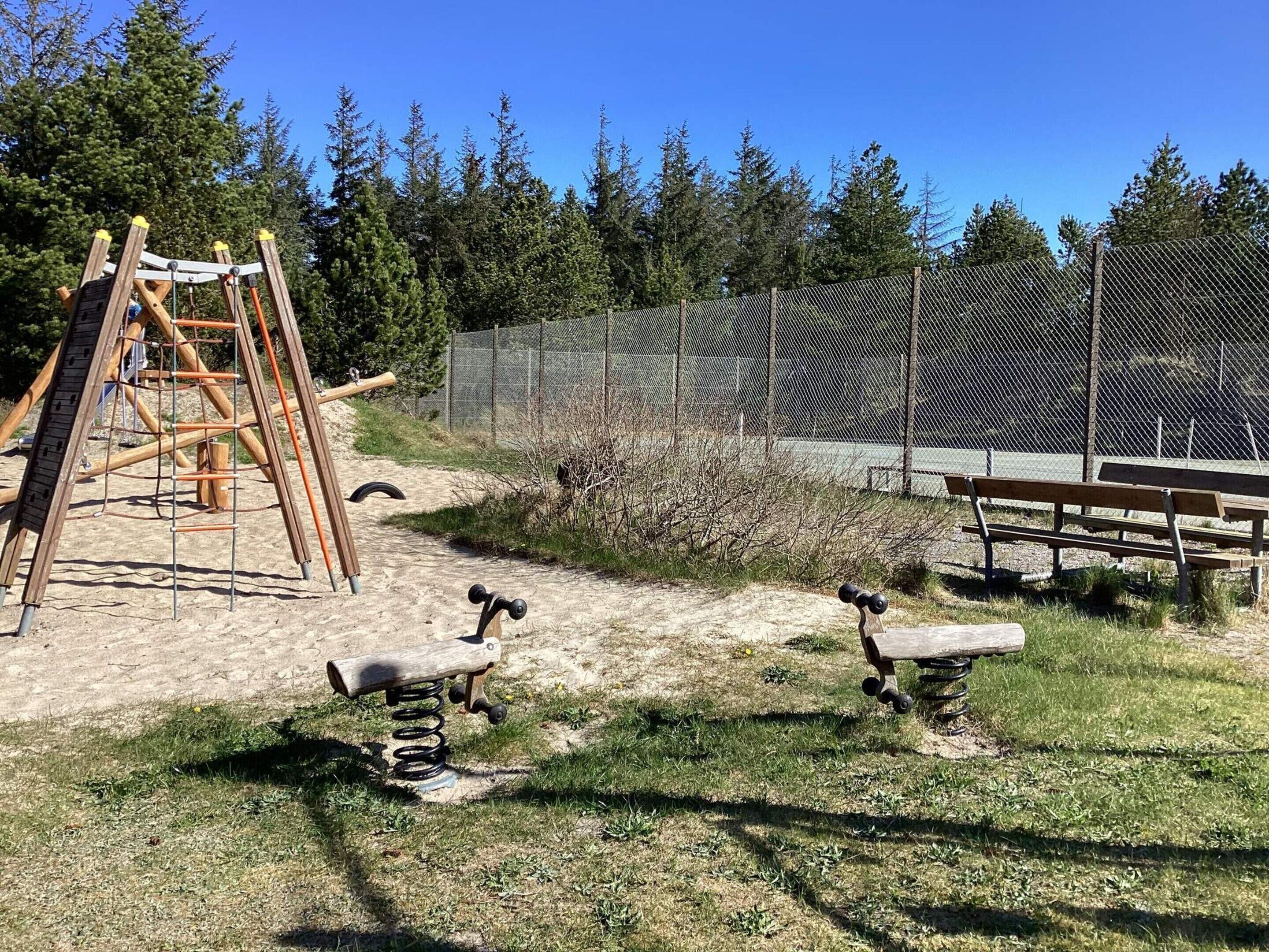 Un parque infantil con un tobogán y un columpio de madera, rodeado por una cerca de malla y árboles.