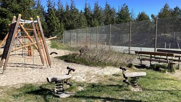 Un parque infantil con un tobogán y un columpio de madera, rodeado por una cerca de malla y árboles.