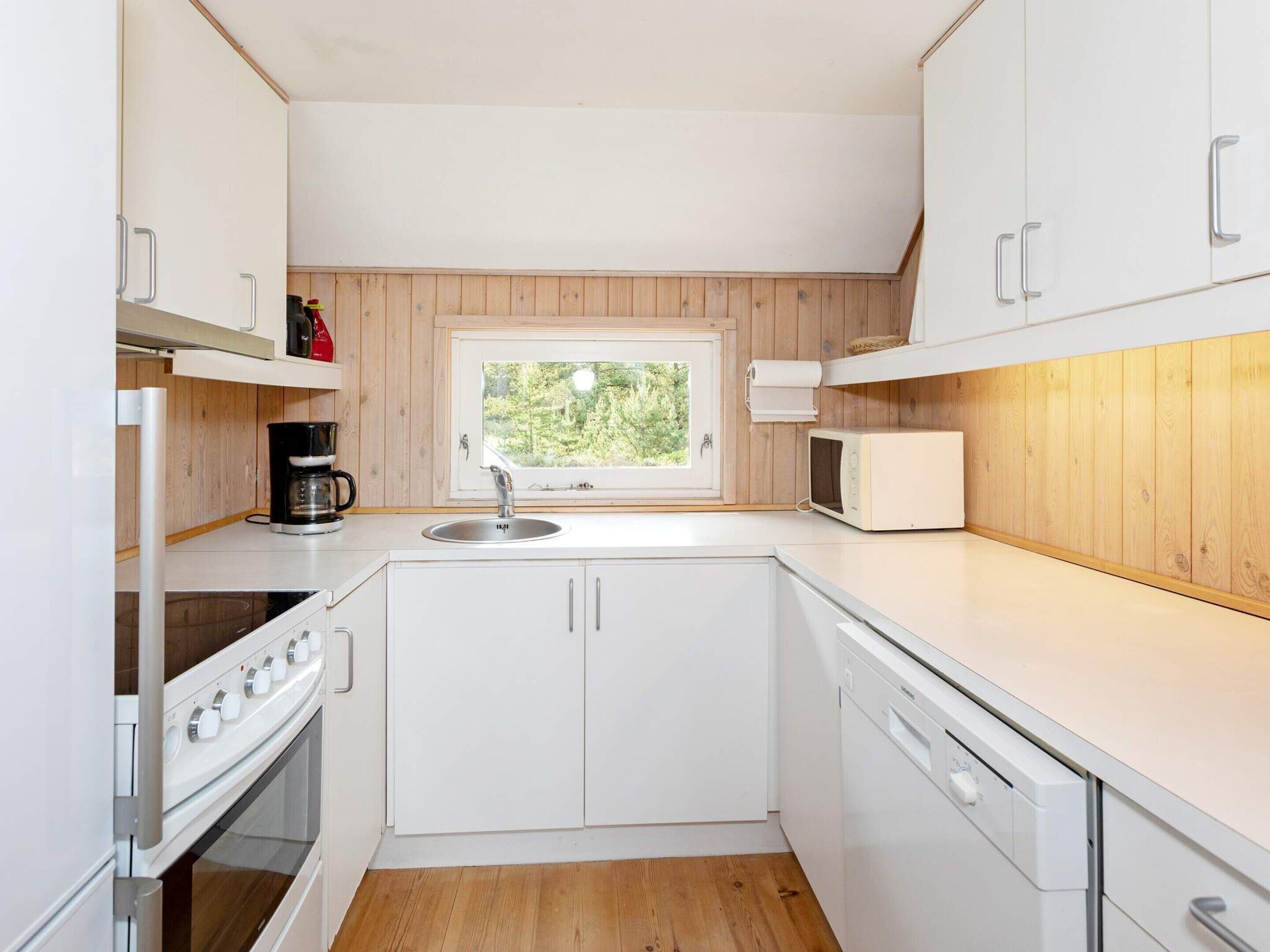 Cocina compacta con armarios blancos, horno integrado, microondas y ventana con vistas al verdor.