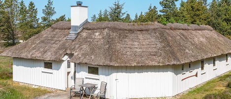 A traditional thatched-roof house with a chimney, a small porch with a table and chairs, and a flag on the side.