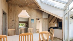 A dining area with a long table and six wooden chairs, a hanging light fixture, and a view through large windows.