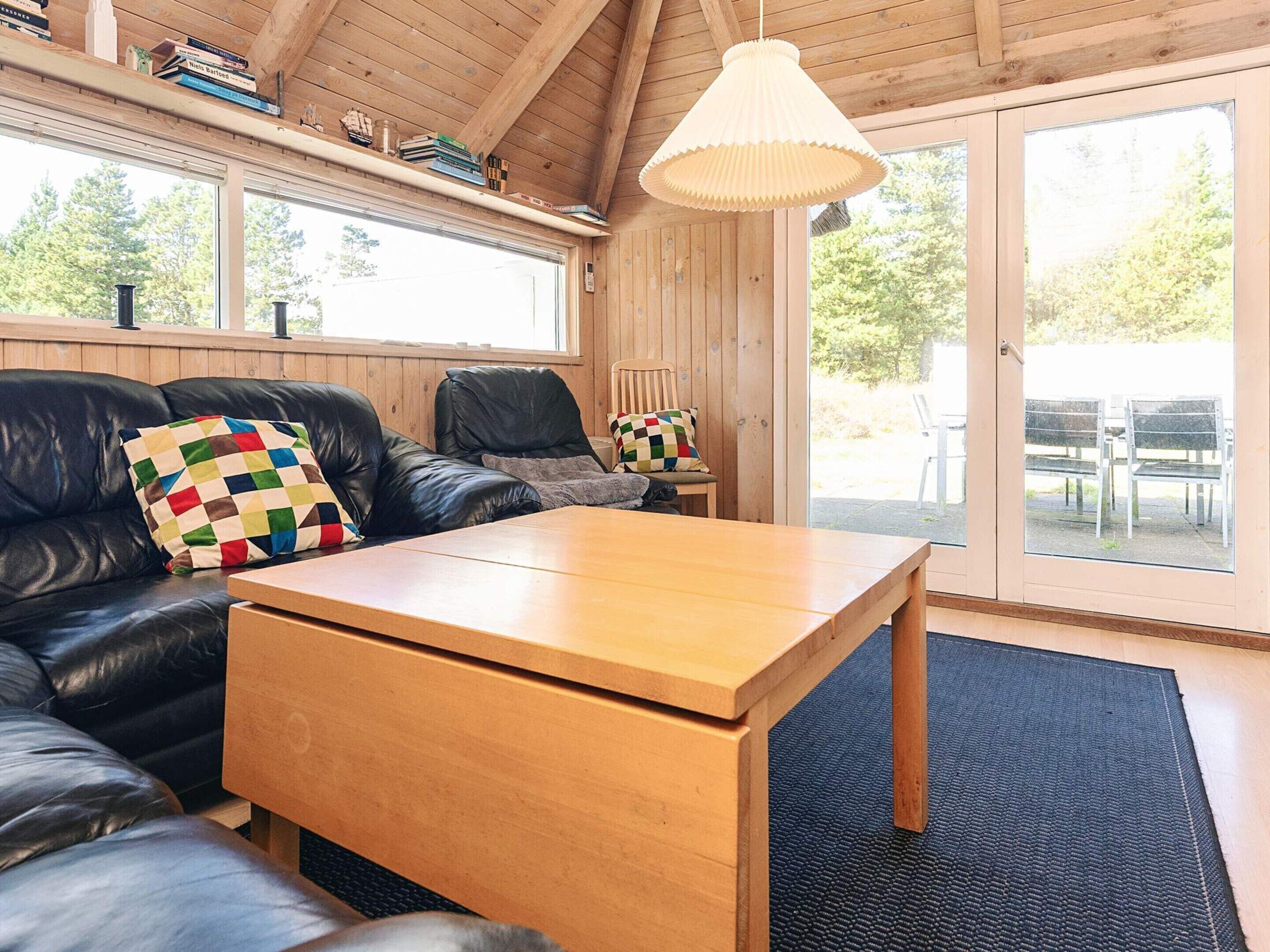 A cozy room with a wooden ceiling, a black leather sofa, a wooden coffee table, and a large window with a view of outdoor seating.