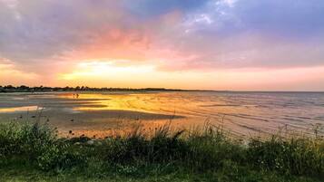 A coastal landscape with a wide sandy beach, grassy areas, and a distant shoreline.