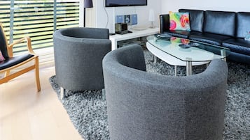 A modern living room with a glass coffee table, two grey armchairs, a black sofa, and a flat-screen TV mounted on the wall.