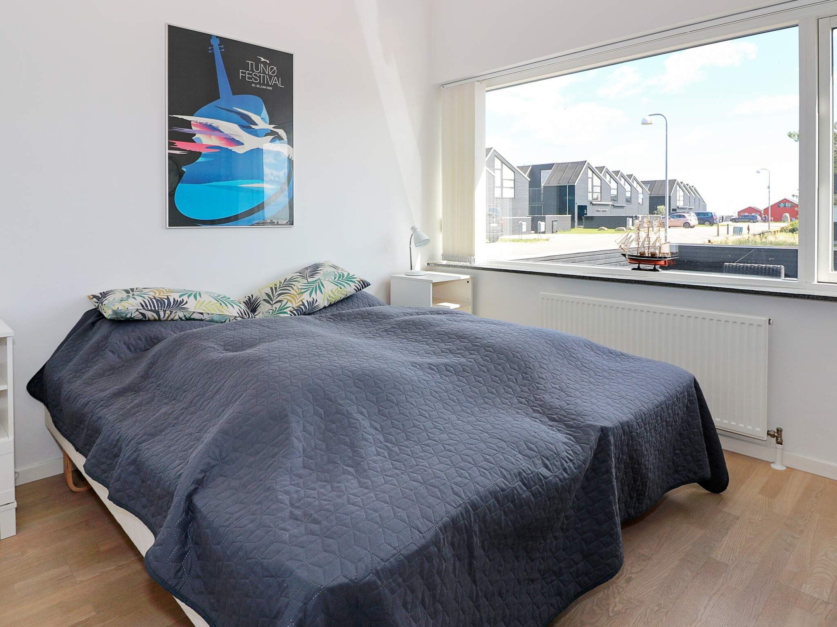 Un dormitorio con cama, ventana con vistas a edificios, un cartel en la pared y una mesita de noche con lámpara.