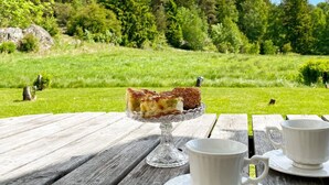 Et træbord med et kagefad af glas, der holder et stykke kage, to hvide kopper på underkopper, og en smuk udsigt over en græsplæne og træer i baggrunden.