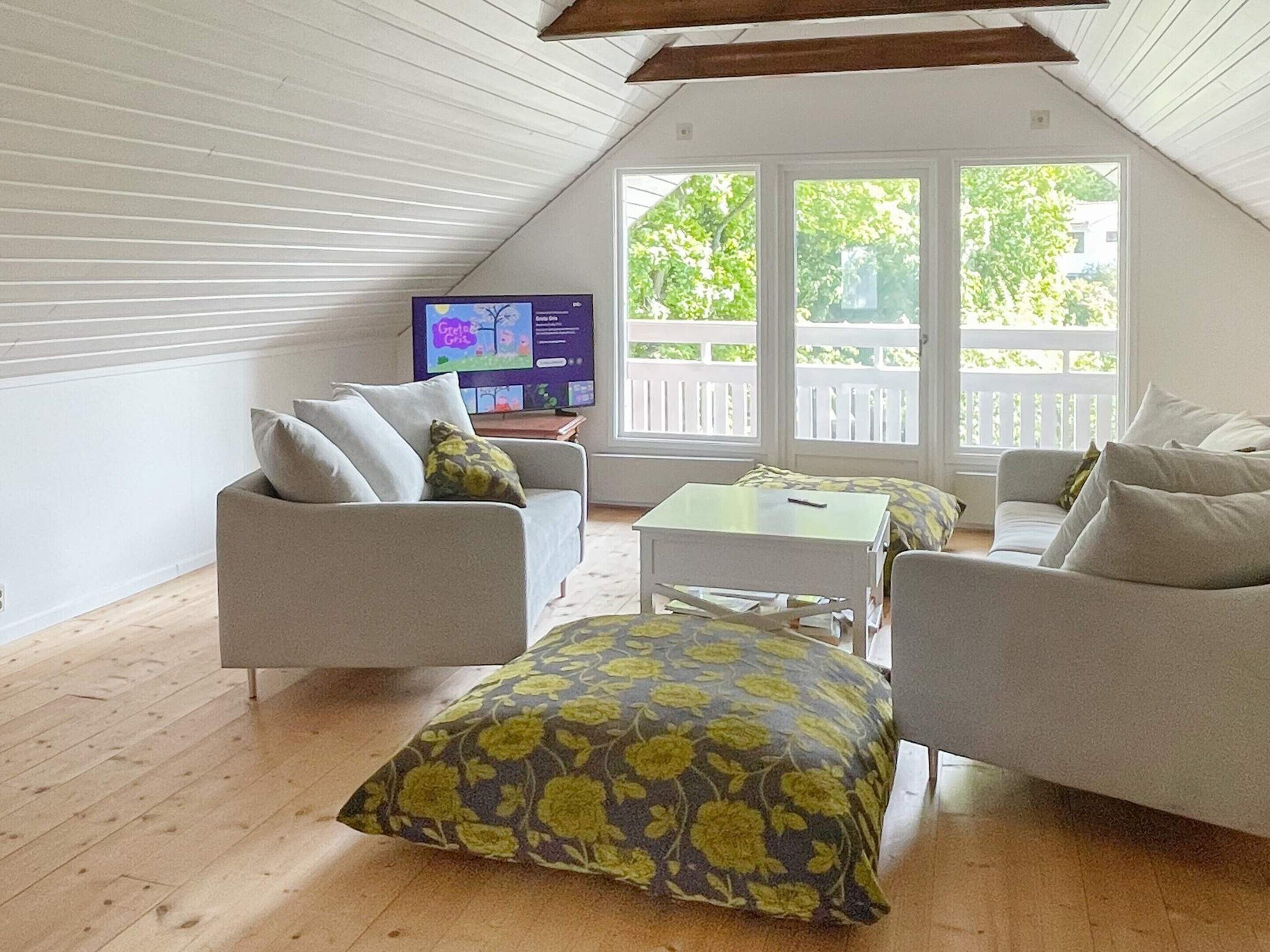 Ett modernt rum i loftstil med ett sluttande tak, ett stort fönster med utsikt över grönområden och en tv-apparat på ett träbord.