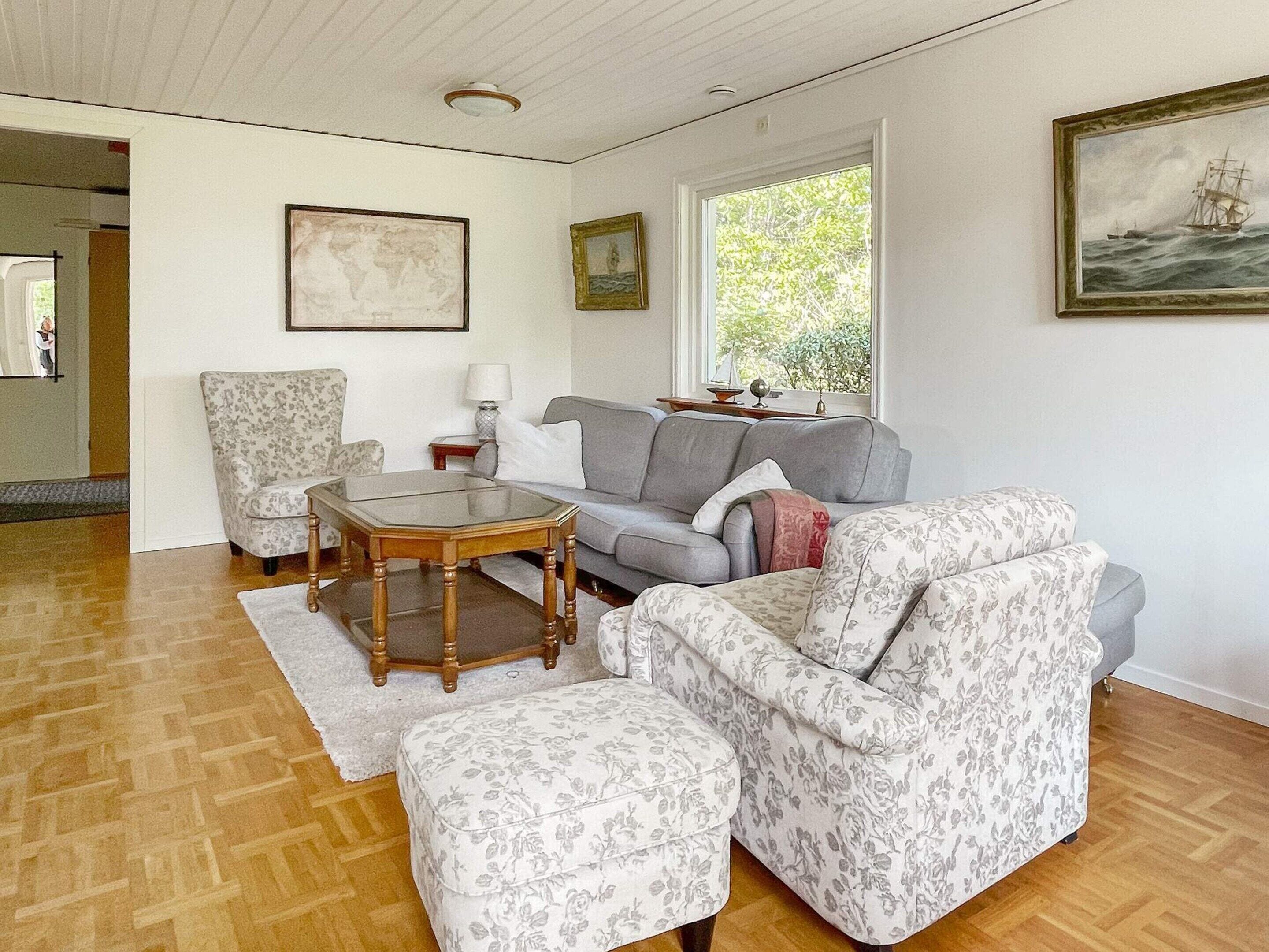 A living room with a sofa, armchair, and ottoman, a glass coffee table, and two framed pictures on the wall.