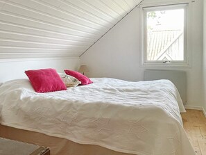 A bedroom with a sloped ceiling, a bed with a white bedspread and two pillows, a nightstand with a lamp, and a window with a view of a white fence. - 4 Star Holiday Home in Branno (Branno)