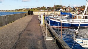 A harbor with boats docked, a concrete walkway, and houses in the background. - 6 Person Holiday Home in Farso (Farso)
