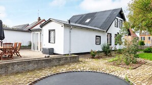 Ein weißes Haus mit schwarzem Dach, eine gepflasterte Terrasse mit Holzstühlen und ein runder Trampolin.