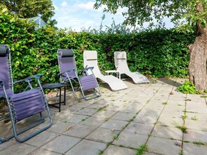 A patio with four lounge chairs and a small table, surrounded by a hedge and a tree.