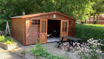 A wooden cabin with a door and windows, a picnic table, and a garden with flowers.