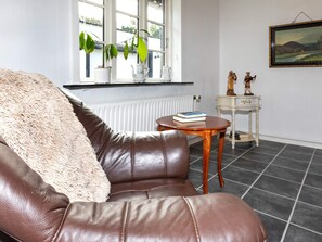 A leather sofa with a fluffy throw, a small round wooden table with a book, a white radiator, and a window with plants.