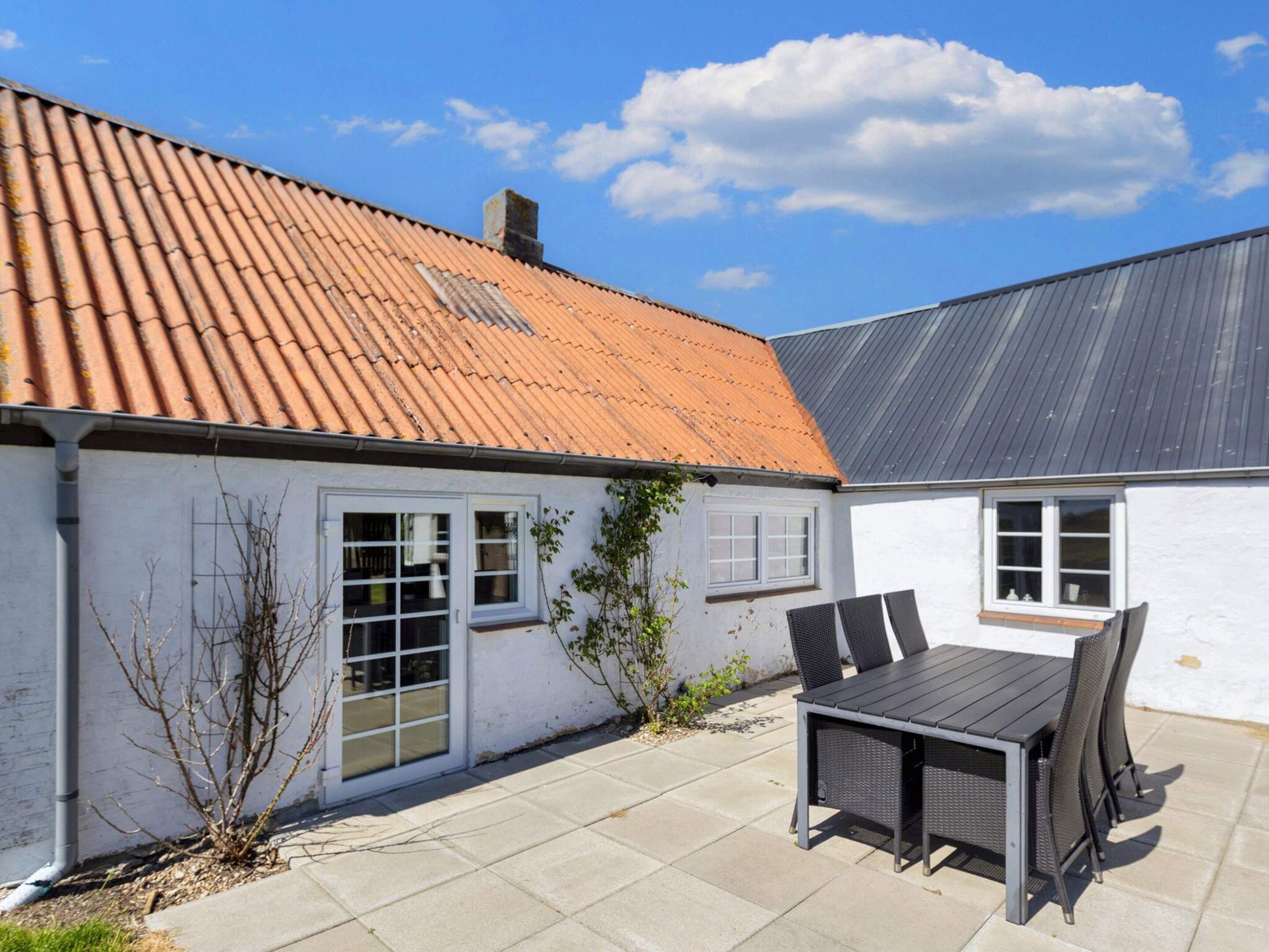 Een huis met een pannendak, een terras met een tafel en stoelen, en een witte buitenmuur.