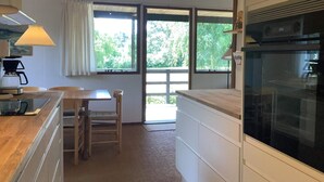 A compact kitchen with a wooden countertop, a dining table, and a chair. There is a window providing a view of greenery outside.