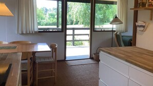 A room with a wooden table, chairs, a lamp, a window with curtains, and a view of greenery outside.