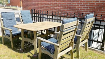 A wooden outdoor table and chairs with blue cushions, set against a brick wall with a metal fence.