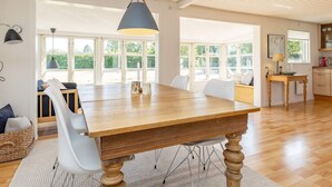 Una habitación amplia con una gran mesa de madera para comer, sillas blancas, una lámpara colgante y ventanas grandes que ofrecen vistas al exterior.