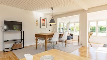 A modern living room with a wooden dining table, white chairs, a TV on a black stand, and artwork on the wall.
