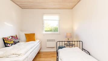 A small, well-lit room with two single beds, a window with a view of greenery, a table with a lamp, and a radiator.