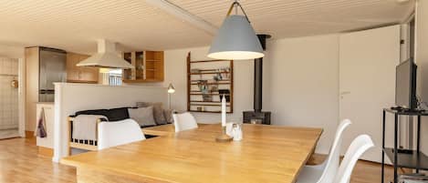 A modern living space with a wooden dining table, white chairs, a kitchen area with cabinets, a built-in bookshelf, and a ceiling with linear lighting.