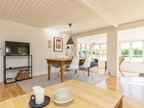 A modern living room with a wooden dining table, white chairs, a television on a black stand, and a large window offering a view of greenery. - 6 Person Holiday Home in Karrebaeksminde (Karrebæksminde)