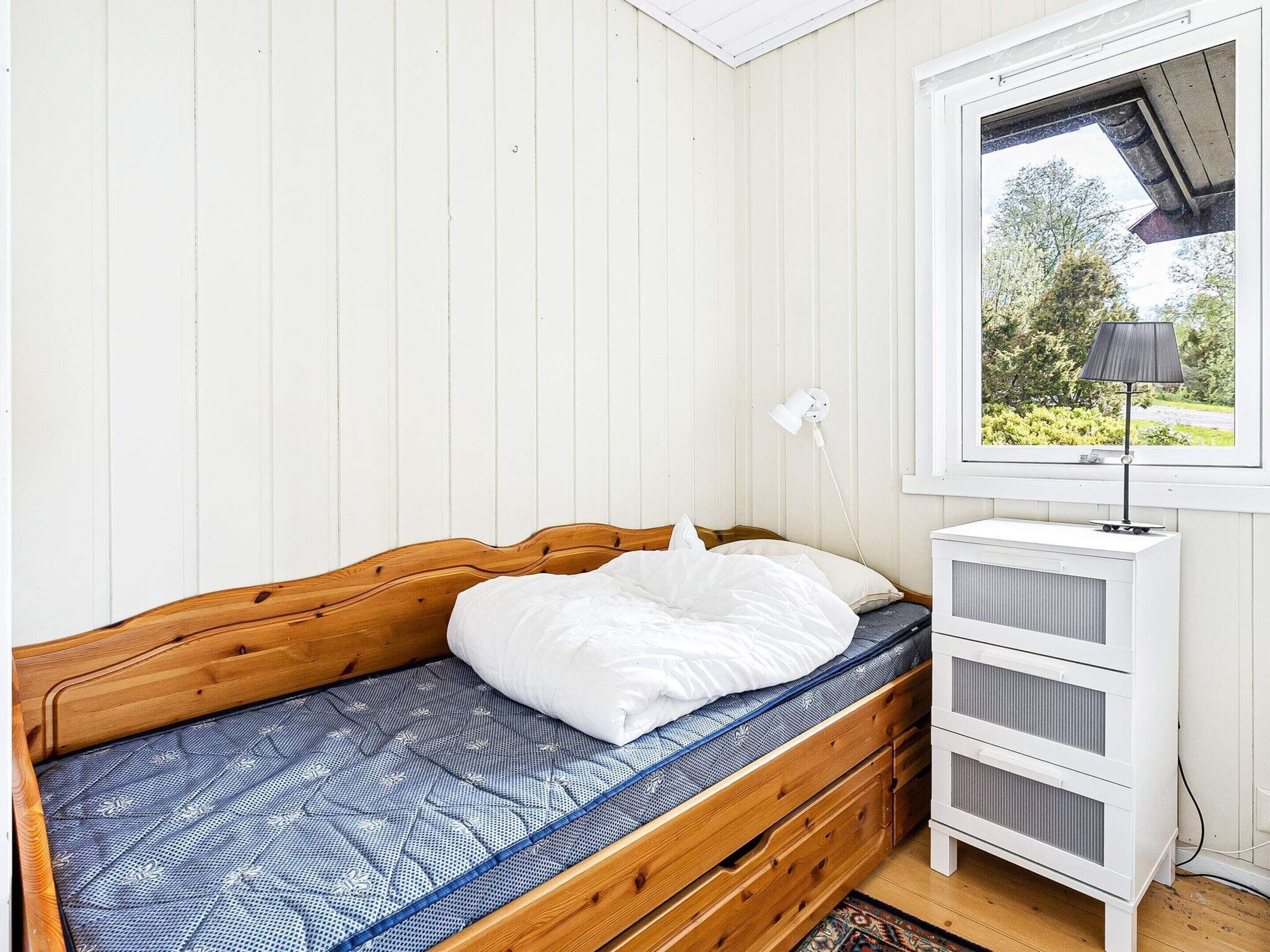 A single bed with a wooden headboard, a white pillow, and a blue patterned mattress cover. There is a white nightstand with drawers and a lamp on it. A window with a view of greenery is visible.