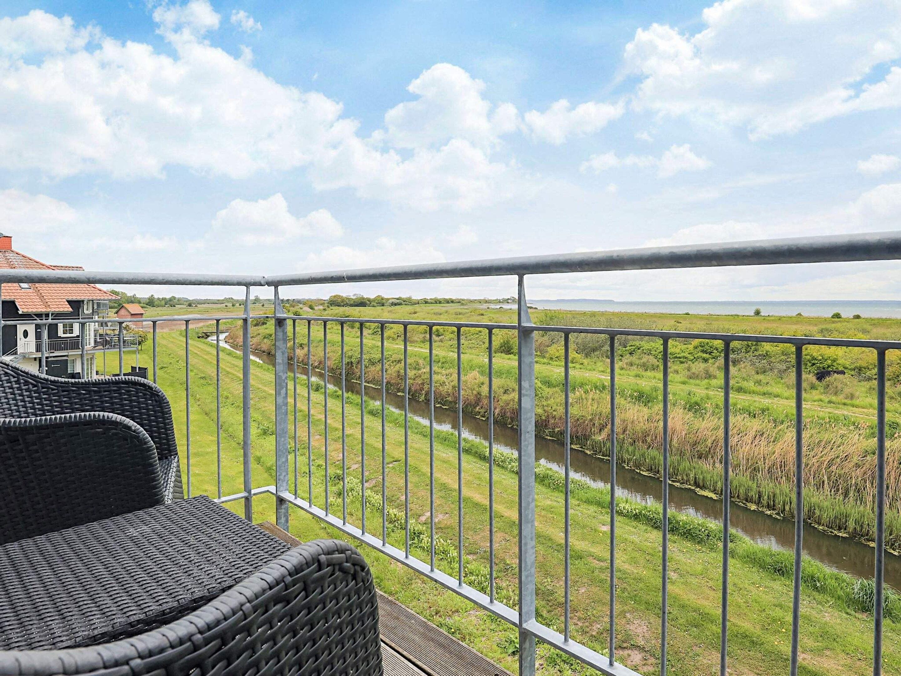 A balcony with a view of a river and greenery, a house in the background, and a clear sky.