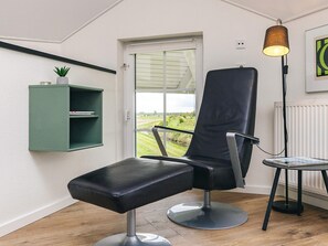 A room with a black leather chair, a small green shelf with a plant, a standing lamp, and a view through a door. - 8 Person Holiday Home in Bogense (Bogense)