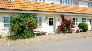 A two-story building with a tiled roof, white exterior walls, and multiple windows. There is a bench and a small garden with red flowers in front. - 6 Person Holiday Home in Sydals (Sydals)