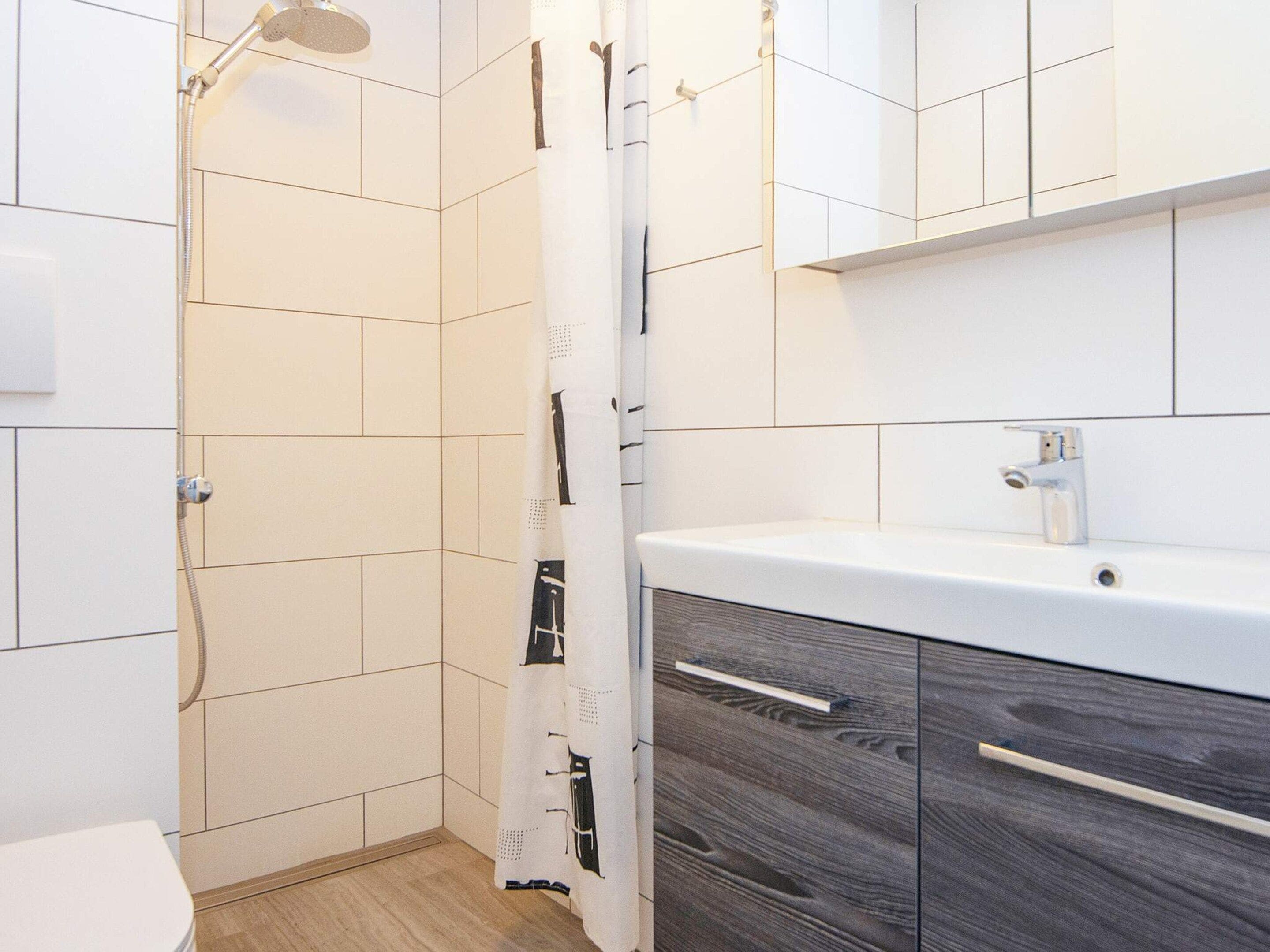 A bathroom with white tiled walls, a shower with a handheld showerhead, a white toilet, and a vanity with a mirror and a sink.