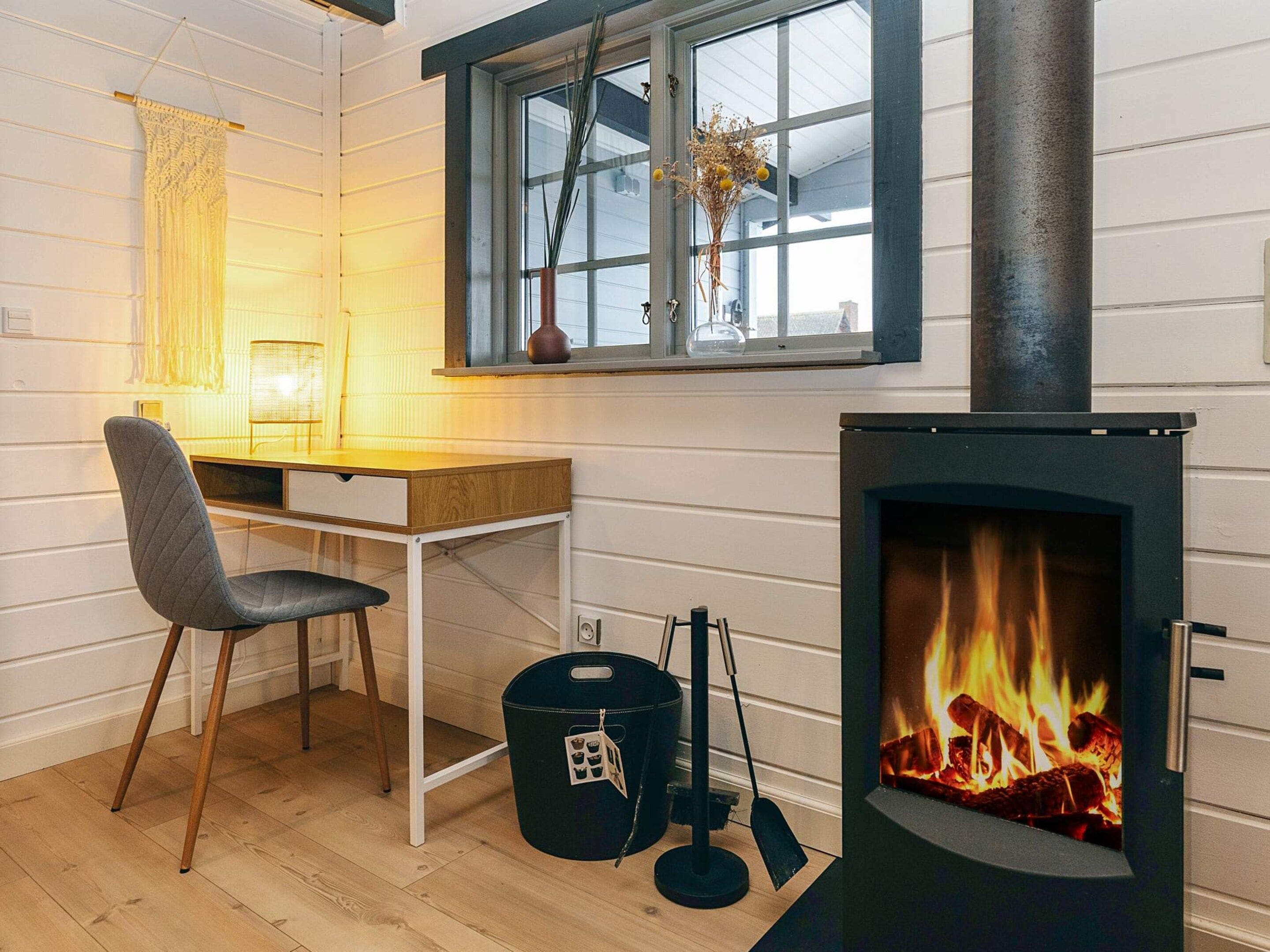 A room with a wooden desk, a chair, a window with a view, and a fireplace with logs.