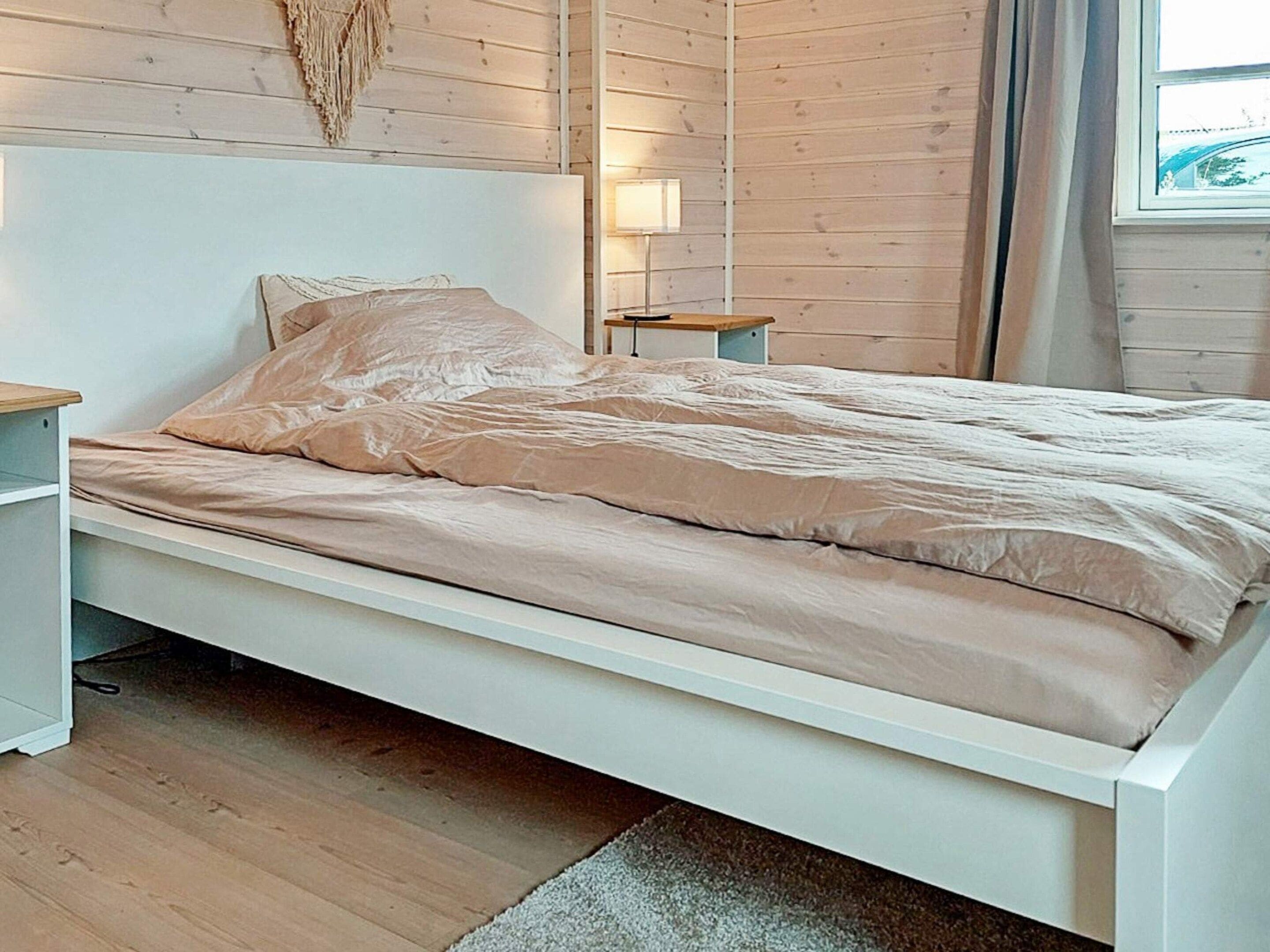 A bed with a white frame and a beige bedspread in a room with wooden walls and a window.