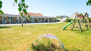 En bygning med rødt tegltag, en overdækket terrasse med borde og stole, og en legeplads med rutchebane og gyngestativer.
