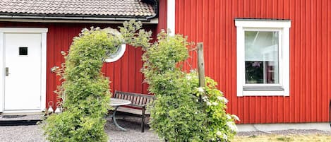 A red building with a white door, a bench, and a small garden area with shrubs.