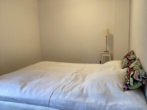 A single bed with a white mattress and pillows, a bedside table with a lamp, and a wall with a plain design. - 7 Person Holiday Home in Motala (Motala)