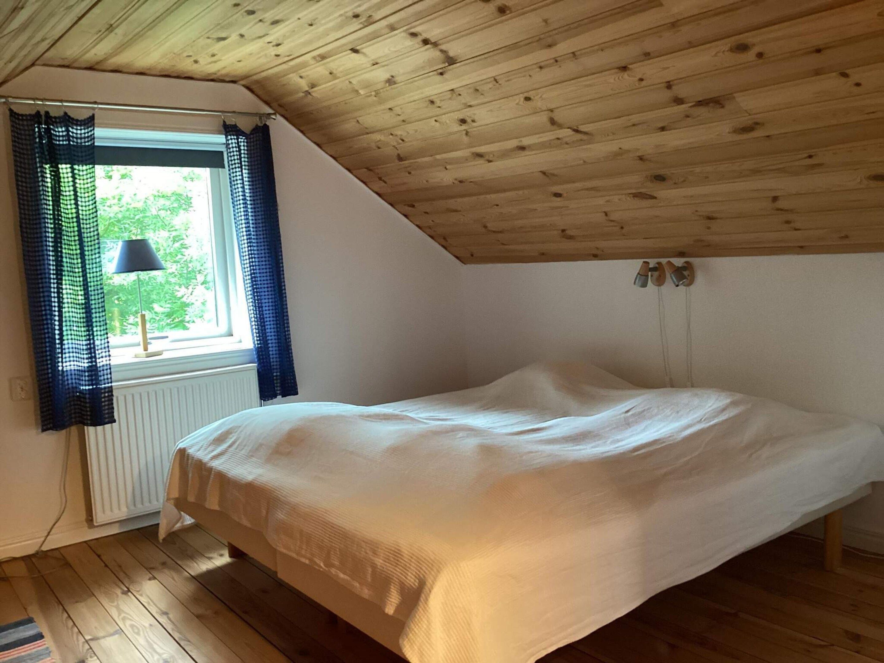 A bedroom with a sloped ceiling, a bed, a window with curtains, and a radiator.