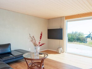 Uma sala de estar moderna com sofá, mesa de centro de vidro, TV na parede e uma grande janela com vista para o exterior.