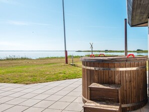 Un bain à remous en bois installé sur une terrasse carrelée, offrant une vue sur un lac et sur la terre ferme au loin.