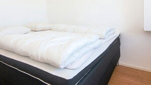 Cama bem arrumada com edredom branco e almofadas, cabeceira preta e piso de madeira.