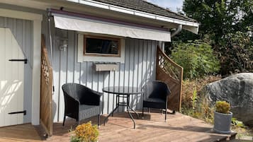 Eine überdachte Terrasse mit zwei Korbstühlen und einem Tisch, einer Holzterrasse und einem Steingarten mit einem kleinen Baum.