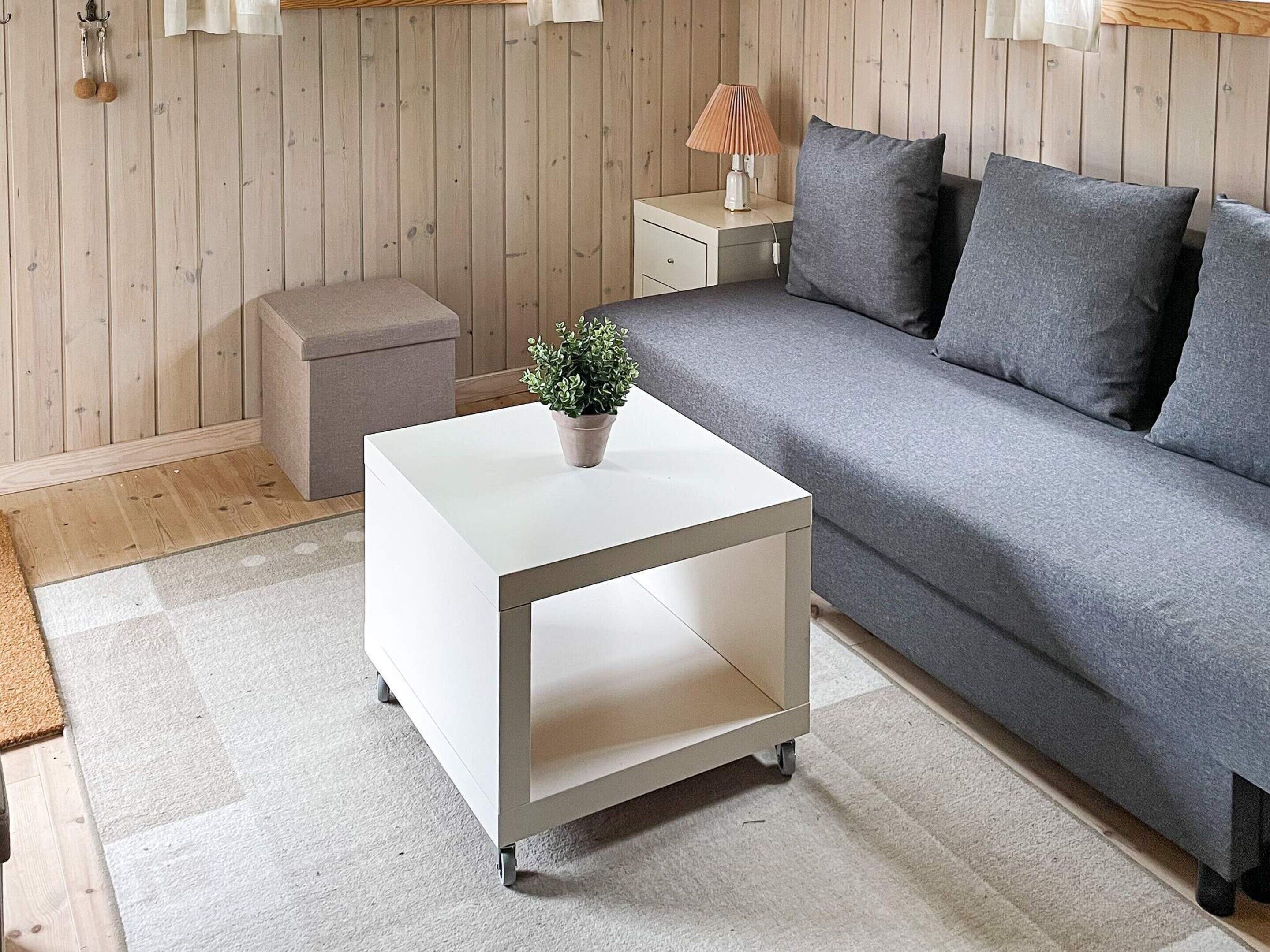 Una habitación con un sofá gris, una mesita de noche blanca con una planta, un pequeño banco de madera y una mesita de noche con una lámpara.