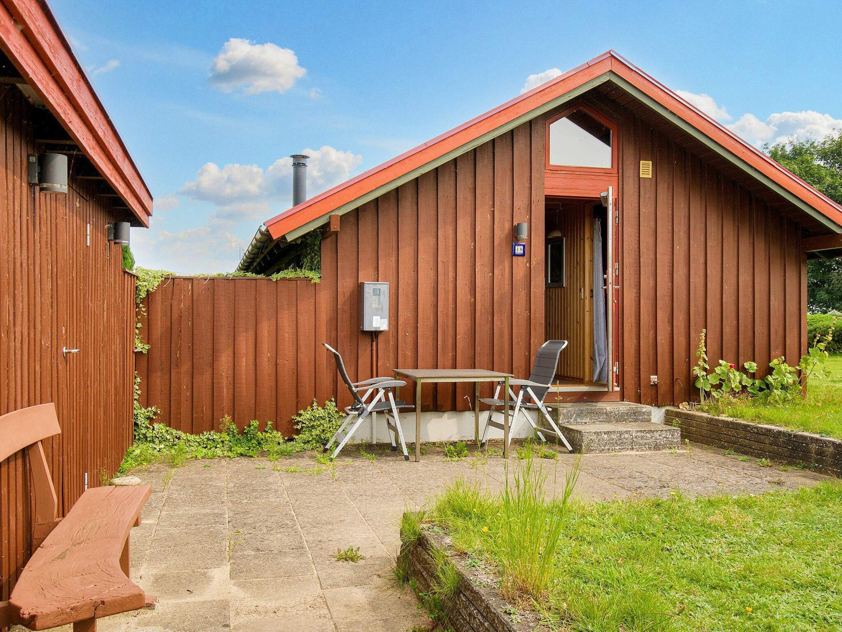 A wooden cabin with a red roof, a small patio with a table and chairs, and a wooden bench.