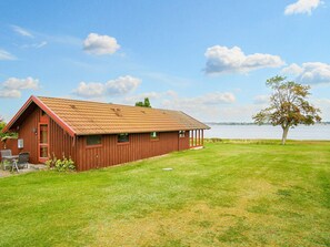 A red wooden building with a sloped roof, a covered porch, and a grassy lawn. - 5 Person Holiday Home in Jaegerspris (Jægerspris)