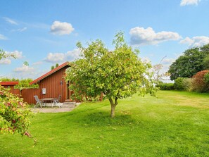 A wooden cabin with a red roof, a small patio with a table and chairs, and a tree with green leaves and red fruits in a grassy yard. - 5 Person Holiday Home in Jaegerspris (Jægerspris)
