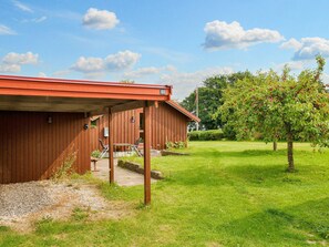 A red wooden building with a covered patio area, a small table and chairs, and a grassy lawn with a tree bearing fruit. - 5 Person Holiday Home in Jaegerspris (Jægerspris)