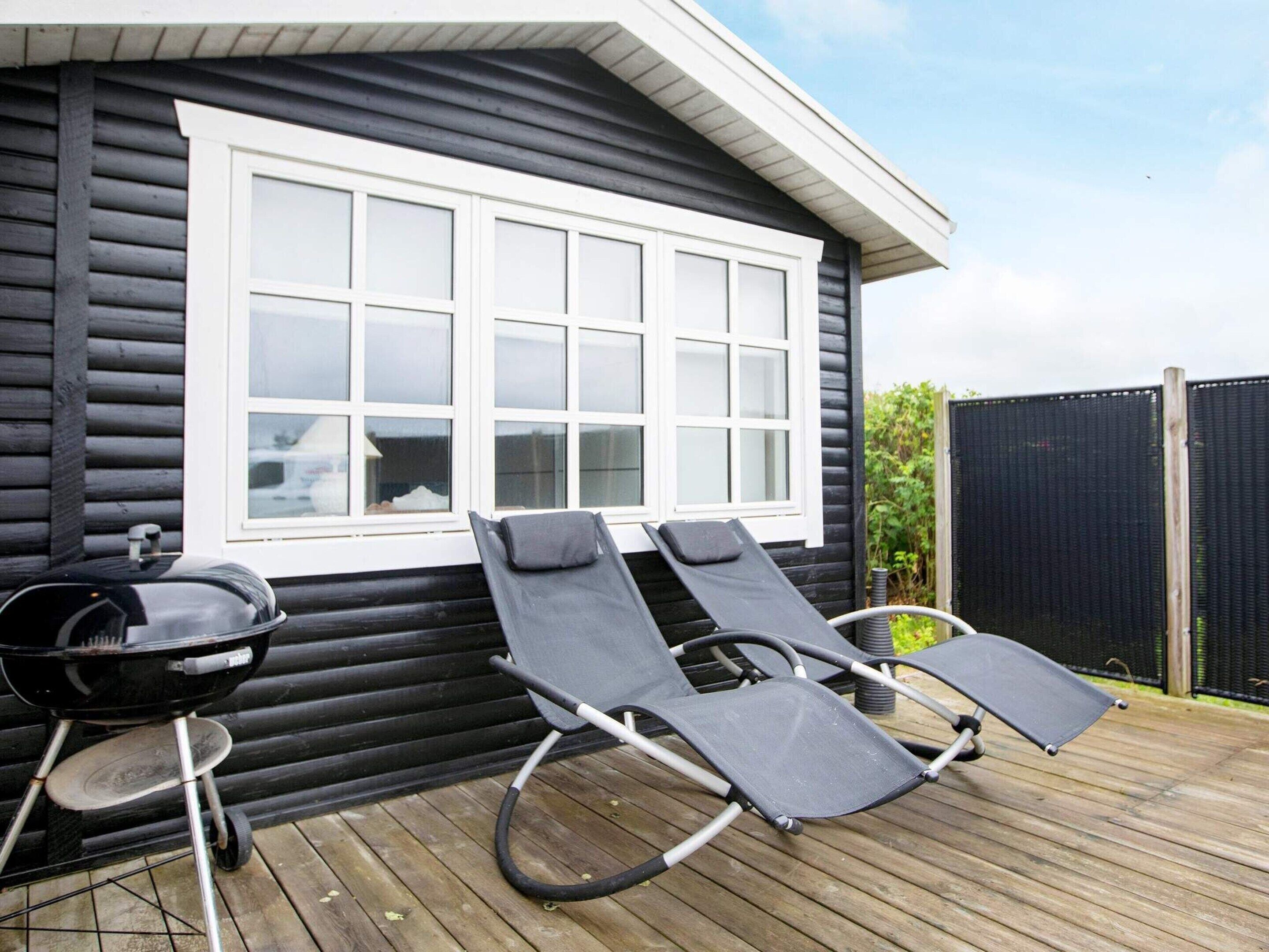 Une cabane avec une terrasse, deux chaises longues et un barbecue.